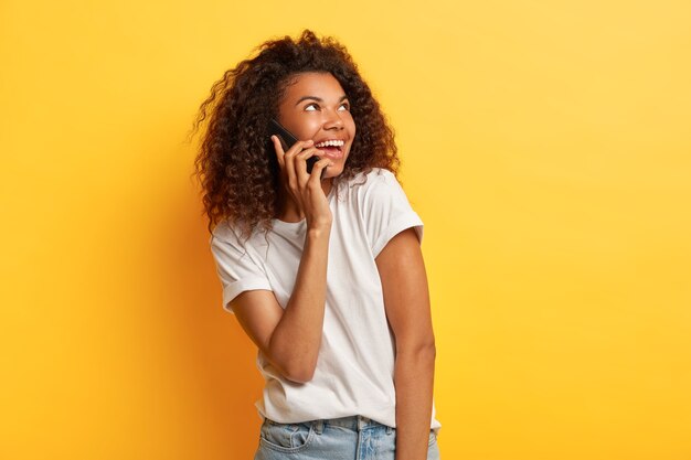 красивая довольная молодая афроамериканка наслаждается приятным разговором, держит мобильный телефон возле уха, смотрит в сторону, носит повседневную белую футболку