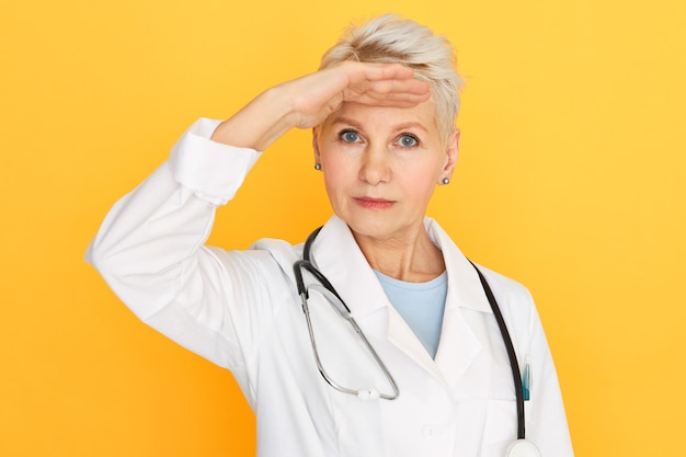 Beautiful senior woman doctor with dyed short hairstyle and blue eyes keeping hand on her forehead searching for something far away in distance.