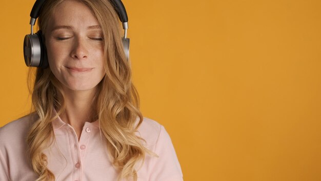 黄色の背景に隔離された目を閉じたままヘッドフォンで新しい曲を聞いている美しい官能的なブロンドの女の子。テキストまたは広告コンテンツ用のスペースをコピーする