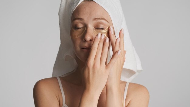 Beautiful sensual woman with towel on head applying eye patches isolated on white background. Beauty concept