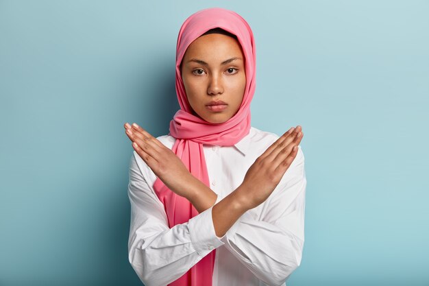 Beautiful serious Arabian woman wears pink hijab, makes cross with hands over chest