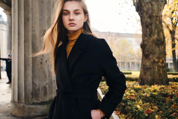 Beautiful serious girl in coat confidently looking in camera in autumn city park