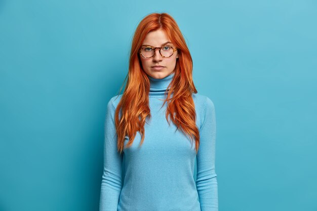 Beautiful serious redhead millennial woman looks directly with calm confident expression listens attentively something wears optical glasses dressed casually.