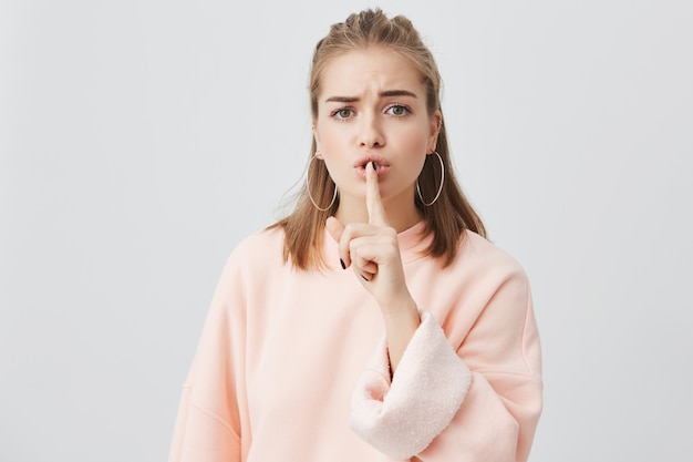 Beautiful serious young female with fair hair dressed in pink sweatshirt and round earings holding finger at lips asking not to make noise and conceal private confidential information. Top secret