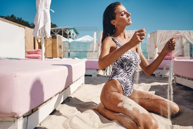 Красивая сексуальная брюнетка с мокрыми волосами в цельном купальнике мечтательно сбрасывает песок с рук на пляж