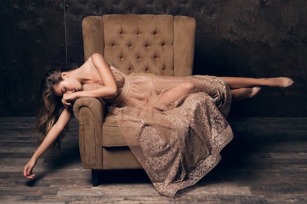 Красивая сексуальная модель женщина в блестящем кружевном вечернем платье позирует, сидя в кресле бежевого цвета