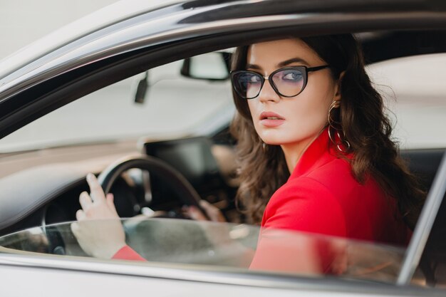 Красивая сексуальная богатая деловая женщина в красном костюме за рулем в белой машине, в очках, стиле бизнес-леди