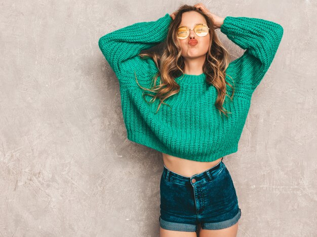 Beautiful sexy smiling gorgeous girl in green trendy sweater. Woman posing in round sunglasses. Model having fun and giving kiss