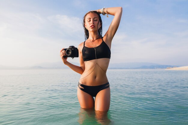 Beautiful sexy woman, perfect slim body, tanned skin, black bikini swimsuit, standing in blue water, holding digital photo camera, hot, tropical summer vacation, fashion trend, waist, belly, hips