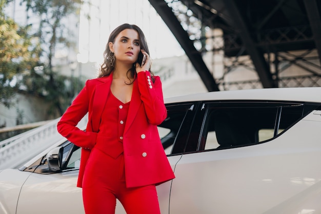 Bella donna sexy in vestito rosso che posa all'automobile che comunica per affari sul telefono