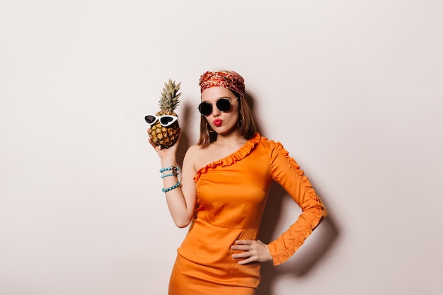 Beautiful short-haired woman in sunglasses and trendy orange dress wore glasses on pineapple.