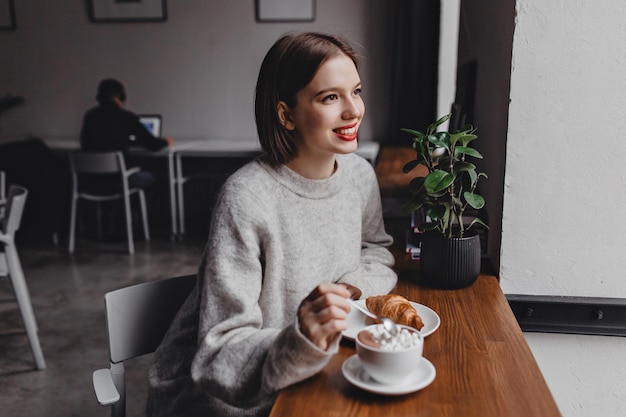 Beautiful shorthaired woman with red lips dressed in gray sweater sits in cafe stirs cappuccino and smiles Picture of girl at table with croissant