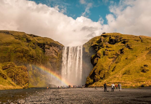 Beautiful shot of people standing near the waterfall with a rainbow on the side in Skogafoss Iceland