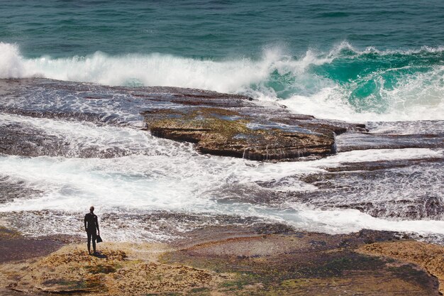 Красивый снимок силуэта человека, стоящего на скале возле пляжа и смотрящего на волны