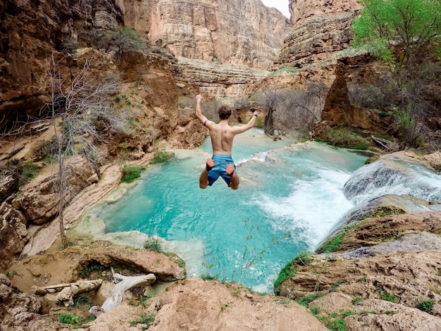 Foto gratuita bello colpo di una persona che indossa il costume da bagno che salta giù da una scogliera in acqua circondata da alberi