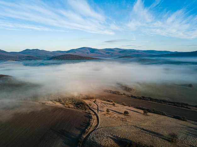 Beautiful shot of a white fog above the field with a road and mountains with a blue sky