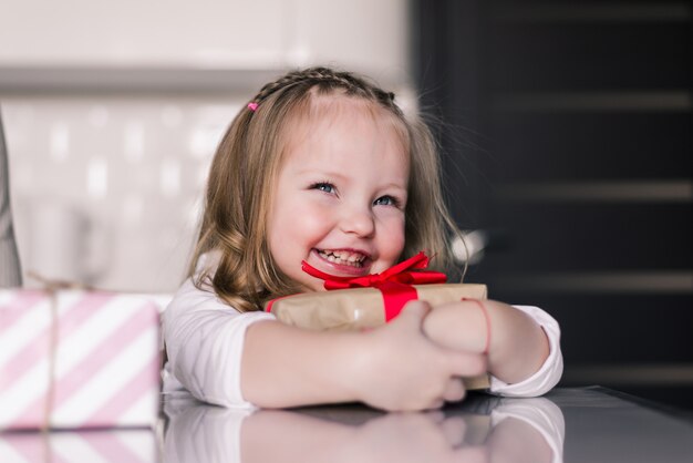 Beautiful small girl holding gifts on her lap and sitting in the kitchen