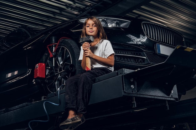 Free photo beautiful small girl is sitting on the lifting platform with car and holding hammer.