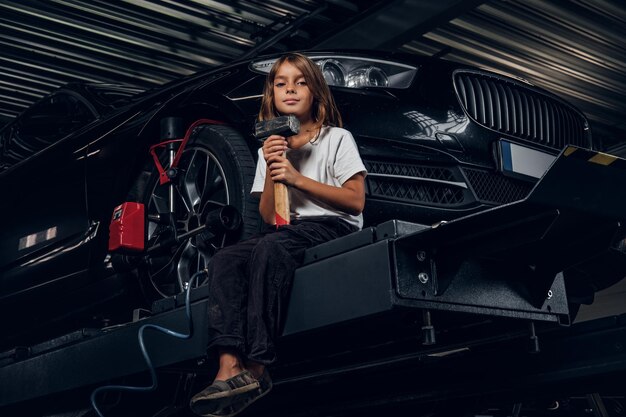 아름 다운 작은 소녀는 자동차와 망치를 들고 리프팅 플랫폼에 앉아 있다.
