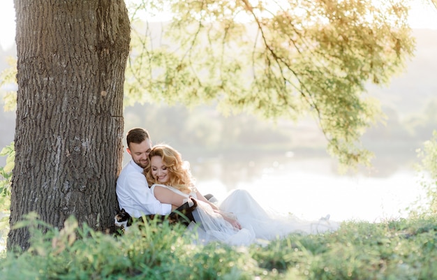 Beautiful smiled couple is sitting on the green grass near tree outdoors, romantic picnic, happy family on the sunny day