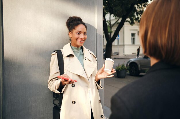 Beautiful smiling African American girl in stylish trench coat emotionally talking with friend outdoor