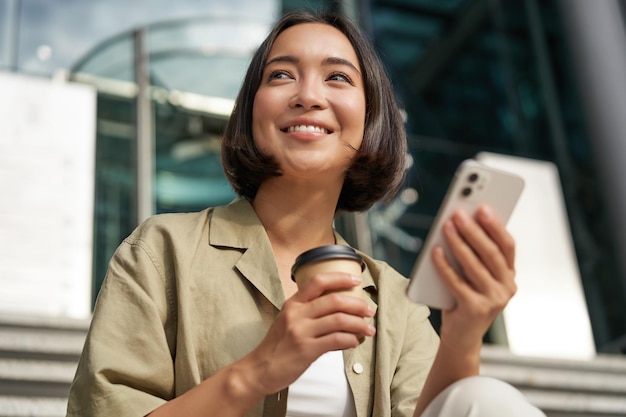 Caffè bevente della bella ragazza asiatica sorridente facendo uso del telefono cellulare e si siede sulle scale fuori del giovane wom