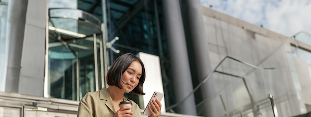 携帯電話を使ってコーヒーを飲み、若い外の階段に座る美しい笑顔のアジアの女の子