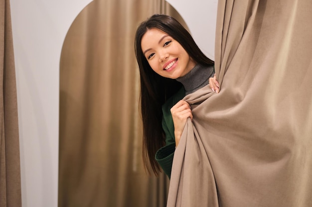 Beautiful smiling Asian girl happily peeping out dressing room in modern fashion store