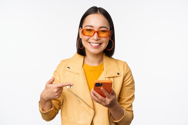 흰색 배경 위에 서 있는 휴대폰에 앱 스토어를 보여주는 스마트폰에서 손가락을 가리키는 선글라스를 끼고 웃고 있는 아름다운 아시아 소녀