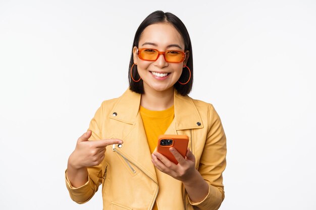 白い背景の上に立っている携帯電話のアプリストアを示すスマートフォンで指を指しているサングラスで美しい笑顔のアジアの女の子