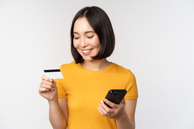 Красивая улыбающаяся азиатская девушка с помощью кредитной карты и мобильного телефона, оплачивающая онлайн на смартфоне, стоящем в желтой футболке на белом фоне