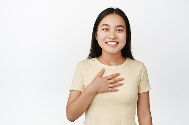 웃고 있는 아름다운 아시아 여성이 가슴에 손을 잡고 흰색 배경 위에 서서 행복해 보이는 자신을 가리키고 있습니다.