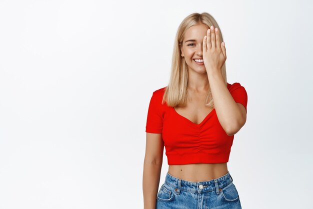 Beautiful smiling blond girl covers half of her face looks with one eye and looks happy at camera stands against white background
