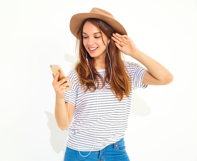 白で隔離されるヘッドフォンで携帯電話で音楽を聴く茶色の帽子でメイクなしのカジュアルな流行に敏感な夏服で美しい笑顔ブルネットの女性女性