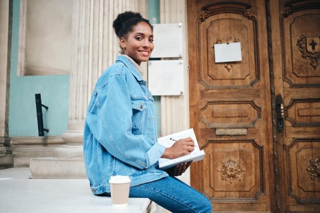 Beautiful smiling casual African American student girl in denim jacket with notebook joyfully looking in camera outdoor