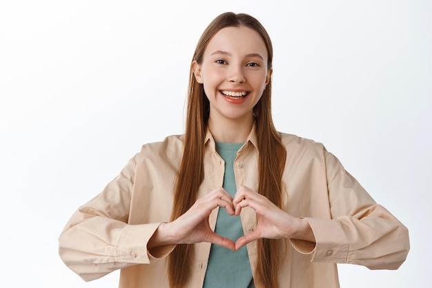 Beautiful smiling caucasian girl shows heart sign I love you gesture care for you standing over white background in casual clothes Copy space