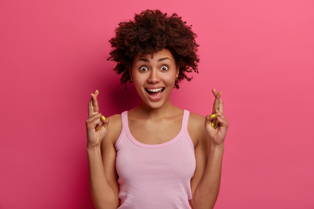 Beautiful smiling dark skinned woman wishes good luck, believes dreams or wish come true, laughs happily, stands with crossed fingers, wears casual clothes, isolated over pink bright wall