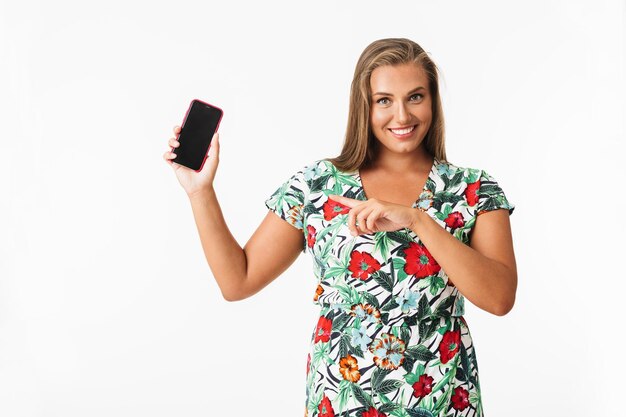 Beautiful smiling girl in colorful dress happily showing new cellphone while looking in camera over white background