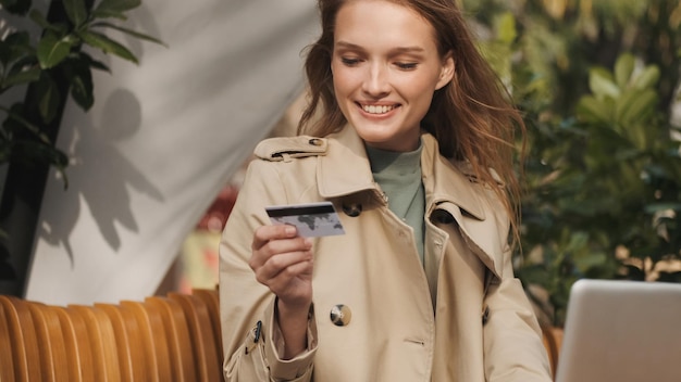 Красивая улыбающаяся девушка, одетая в плащ, держит кредитную карту и улыбается, работая на ноутбуке в кафе на открытом воздухе Современные технологии