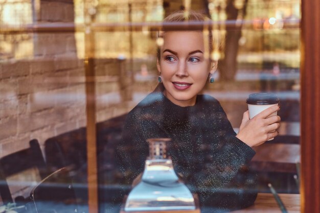 A beautiful smiling girl drinking coffee in the cafe behind the window.