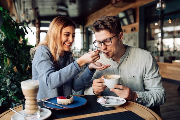 Beautiful smiling girl feeds her handsome boyfriend, eating tasty cake and drinking coffee