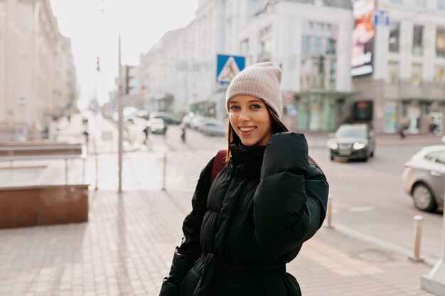 Beautiful smiling girl in hat and jacket is looking at camera on city center in sunlight