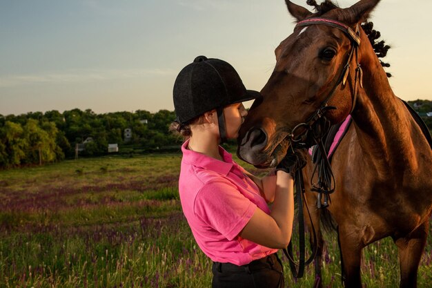 空に特別な制服を着て、夕日に緑の野原の背景を身に着けている彼女の茶色の馬の横に立っている美しい笑顔の女の子の騎手。