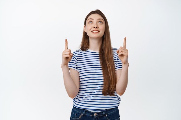 Free photo beautiful smiling girl looking, pointing fingers up with pleased dreamy face, white background.