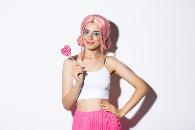 Красивая улыбающаяся девушка в розовом парике, держащая конфету в форме сердца, трюк или угощение в костюме феи на хэллоуин, стоя.