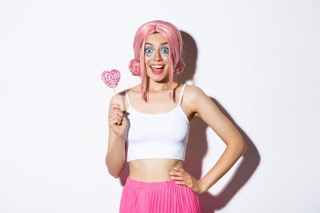 Free photo beautiful smiling girl in pink wig, holding heart-shaped candy, trick or treating in fairy costume on halloween, standing.