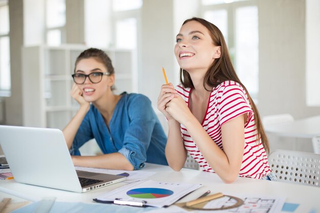 Beautiful smiling girl in striped Tshirt happily looking aside while holding pencil in hand with bored girl near Young women working with laptop in modern office together