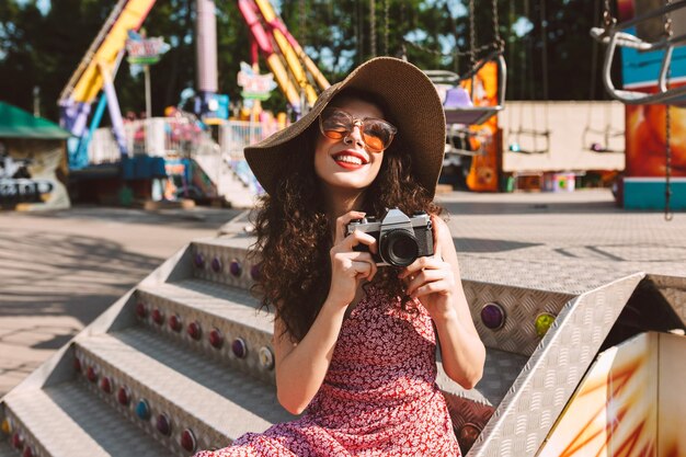 Beautiful smiling girl with dark curly hair in sunglasses and hat sitting with little camera in hand while happily spending time in amusement park