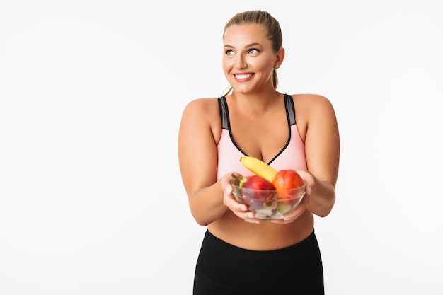 Beautiful smiling girl with excess weight in sporty top holding bowl with fruits in hands while happily looking aside over white background