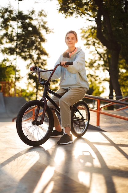 Beautiful smiling girl with headphones on bicycle dreamily looking in camera at modern skatepark